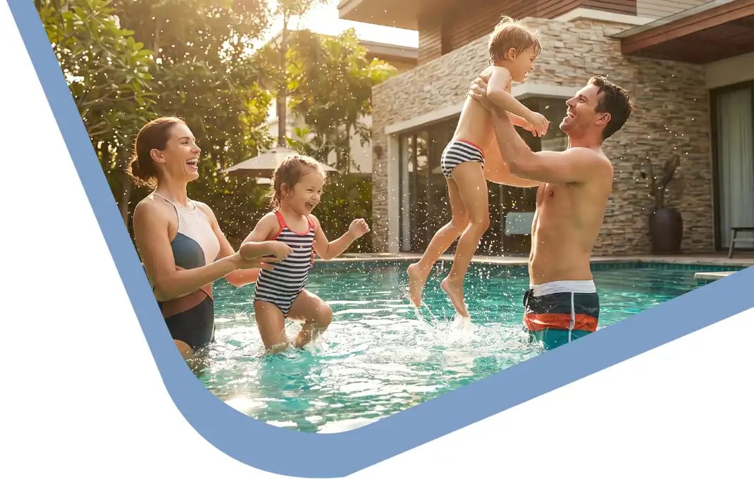 Safe Snacking by the Pool- How to Avoid Allergen Exposure Outdoors-optimized A happy family playing together in an outdoor swimming pool on a sunny day in front of a modern home. A father lifts a young boy playfully into the air while a mother smiles and interacts with a young girl in the water, splashing happily.